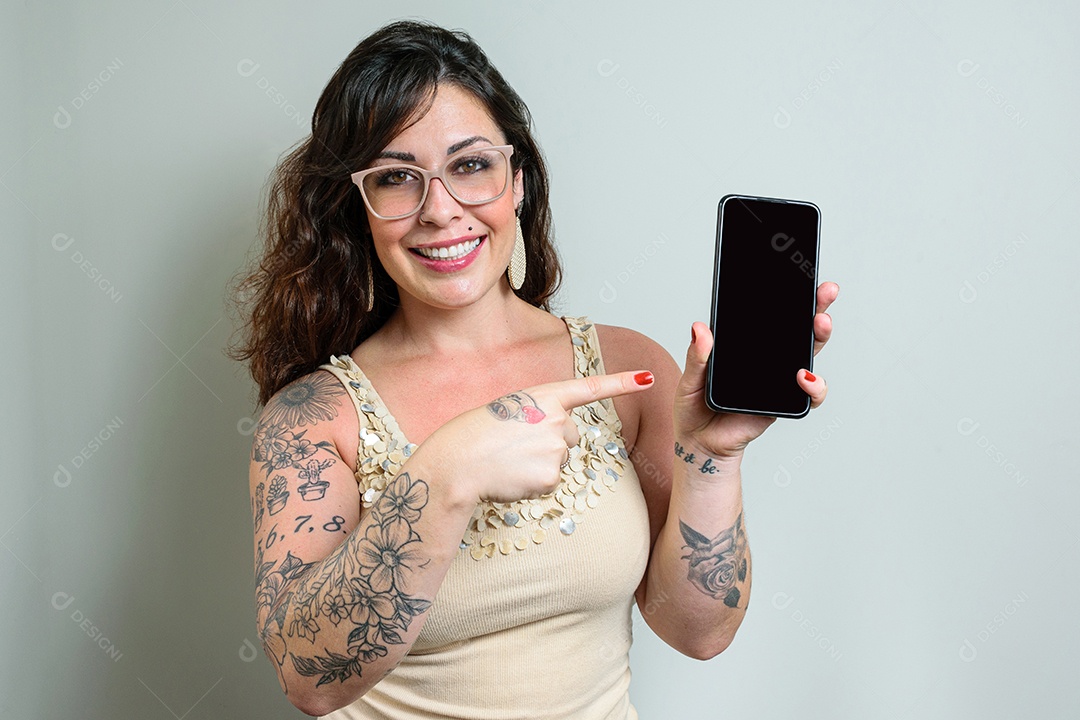 Mulher brasileira de óculos, tatuada, sorrindo e apontando o dedo para o smatphone