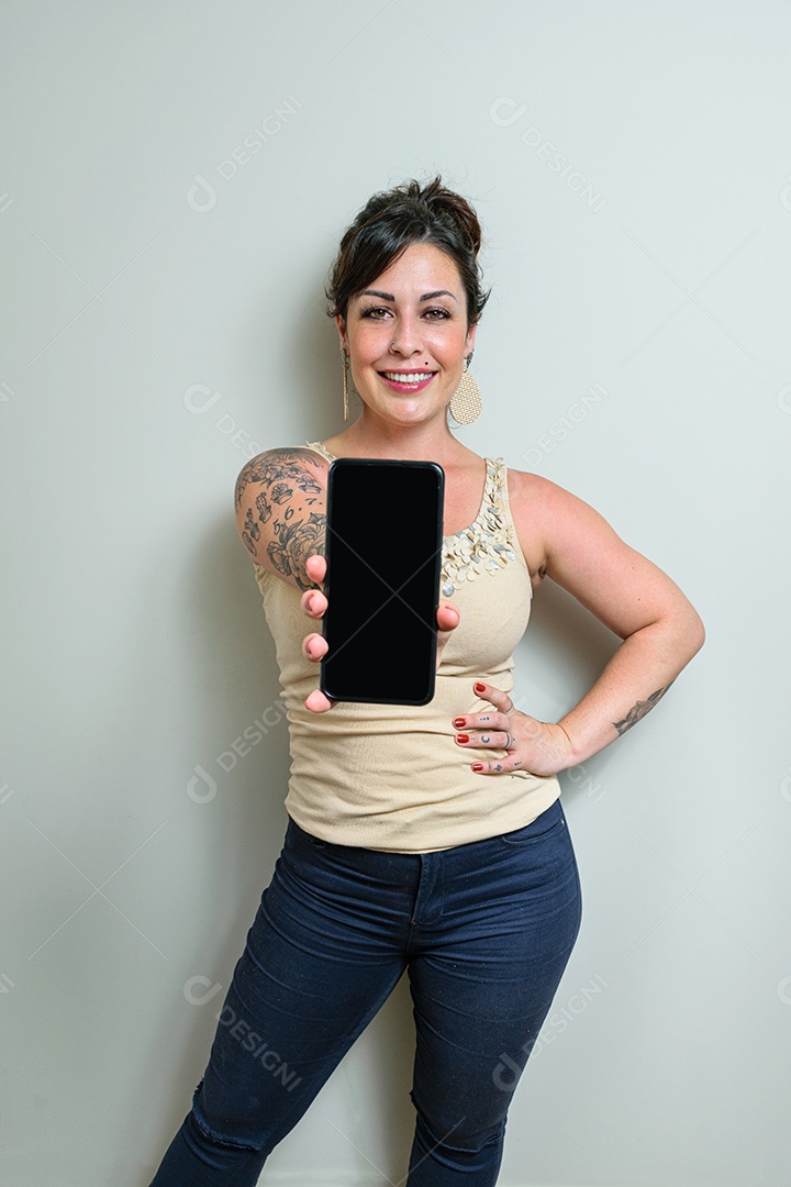 Mulher brasileira, tatuada, cabelo preso, sorrindo e mostrando smartphone