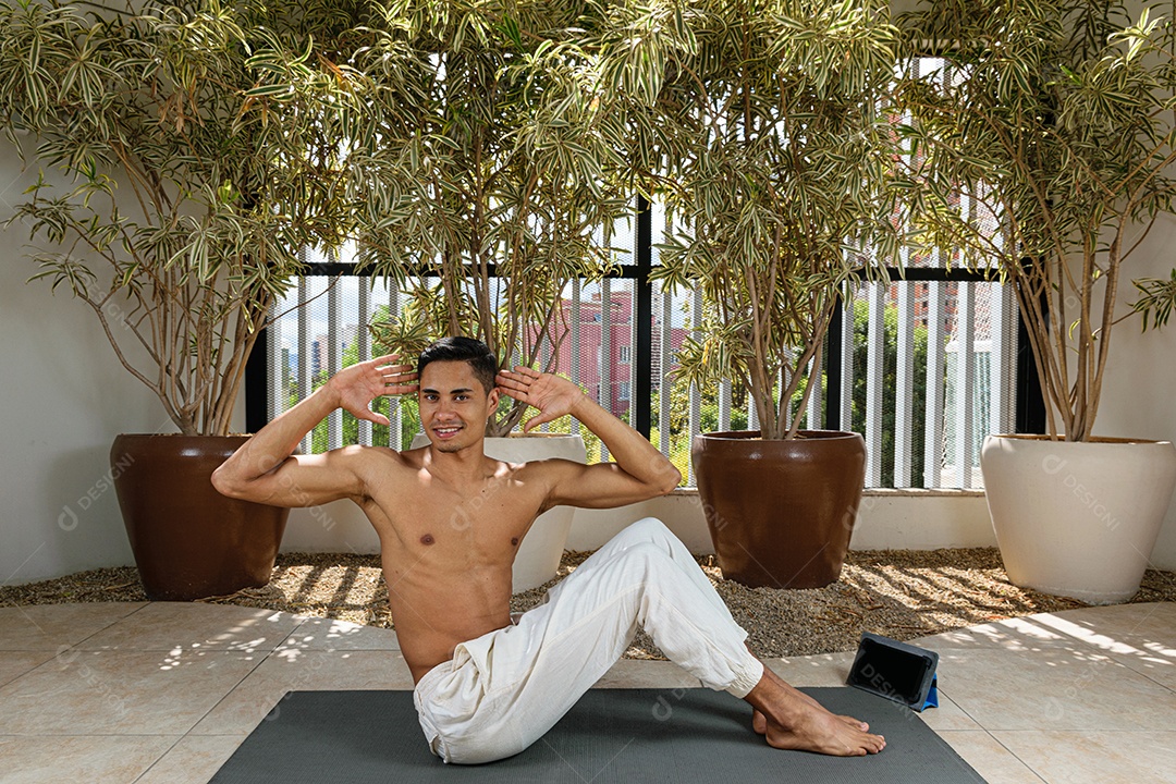 Jovem brasileiro sorridente fazendo aula de ioga on-line, com as mãos na cabeça