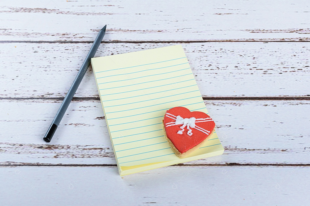 Bloco de notas ao lado de um lápis grafite e um biscoito em forma de coração.
