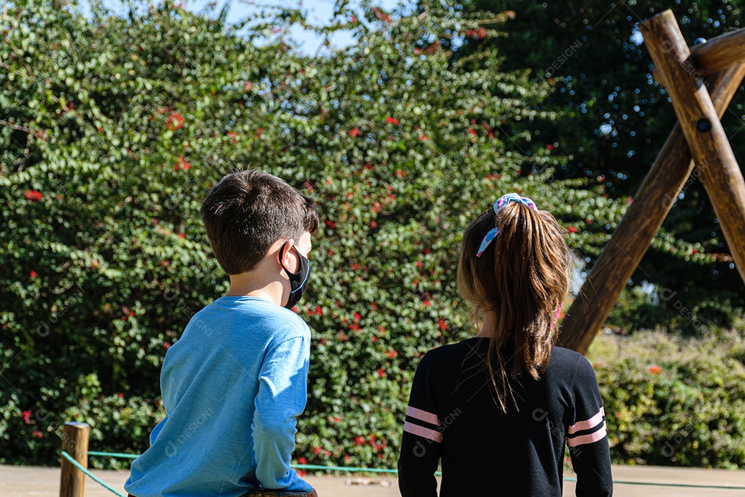 Um menino e uma menina de costas e usando máscara, assistindo a um balanço.