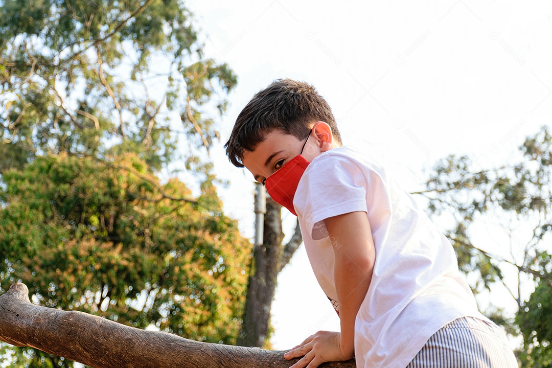 Criança de 8 anos, com máscara, em tronco de árvore em tarde ensolarada