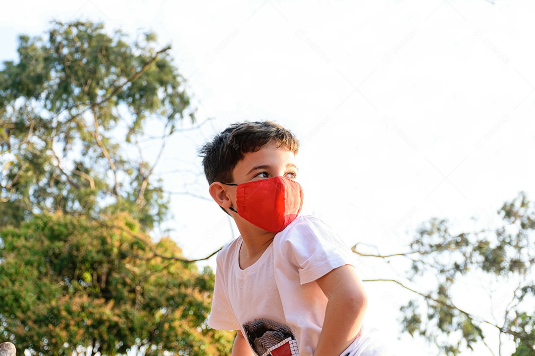 Criança de 8 anos, com máscara, olhando para o lado em uma tarde ensolarada