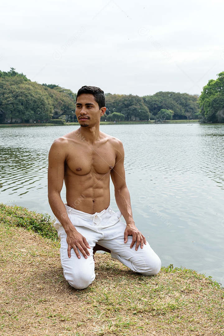 Jovem brasileiro sentado de joelhos à beira de um lago, sem camisa