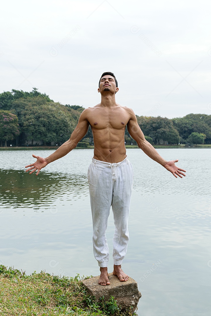 Jovem brasileiro em pé ao lado de um lago, sem camisa, com os braços abertos
