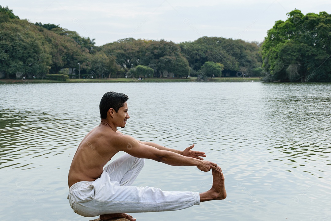 Jovem brasileiro agachado à beira de um lago, sem camisa, alongando-se