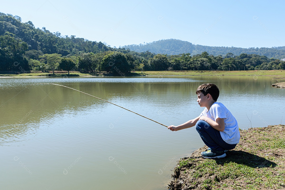 Criança brasileira de 8 anos, agachada à beira do lago, pescando