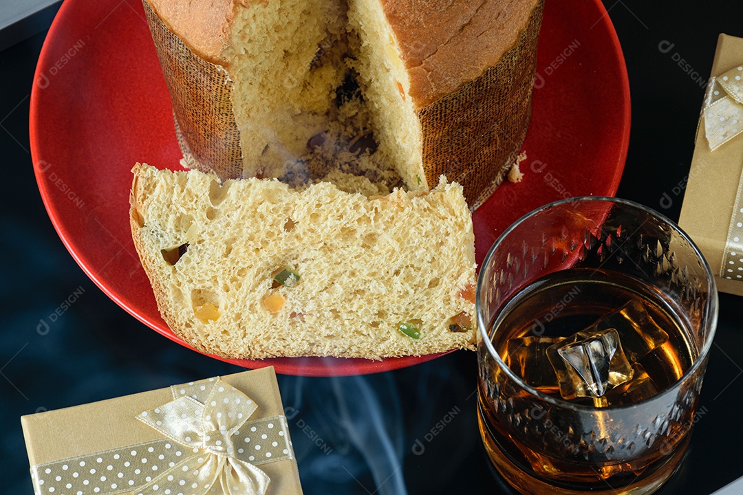 Fatia de panetone sob fumaça de cigarro, ao lado de uma caixa de presente e um copo de uísque virado.