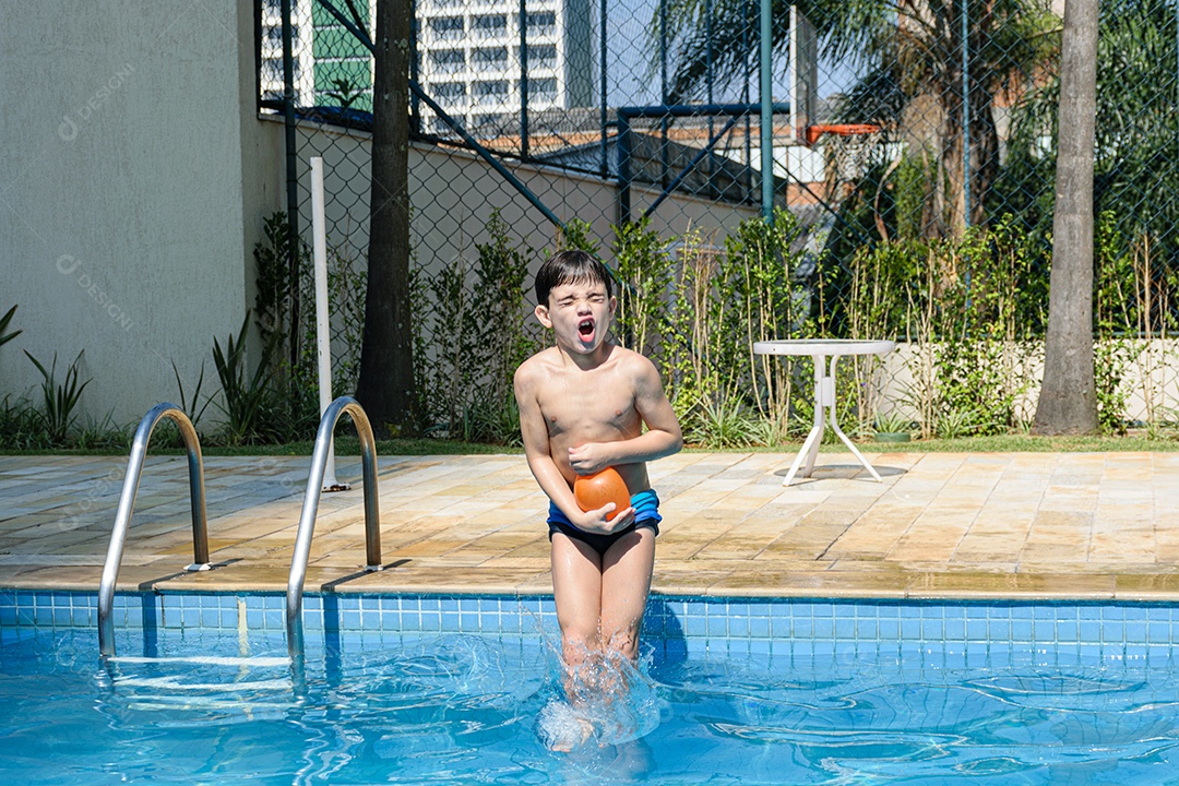 Criança de 8 anos fazendo careta e pulando na piscina com uma bomba dágua.