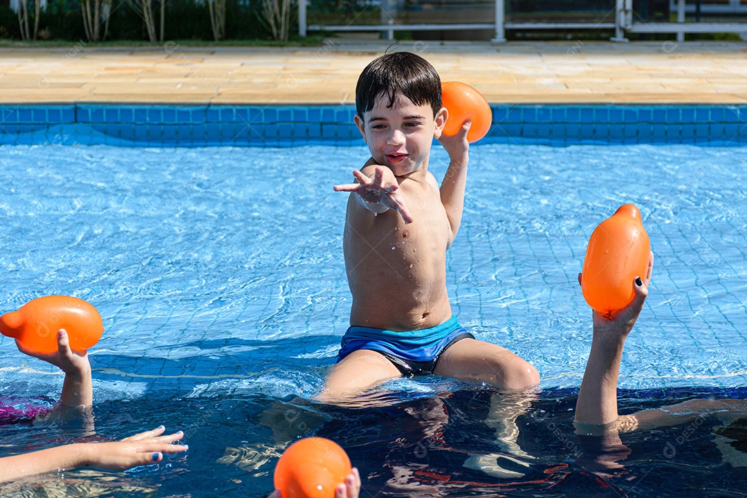 Criança de 8 anos sentada na beira da piscina com a boca aberta e cercada por outras mãos com bombas dágua.