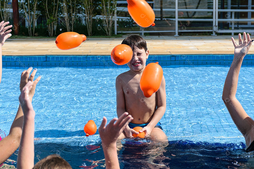 Criança de 8 anos sentada na beira da piscina, brincando com várias bombas dágua.