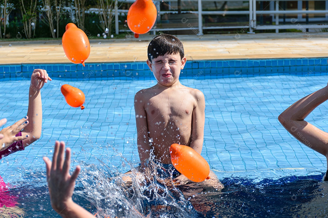 Criança de 8 anos sentada na beira da piscina, brincando com várias bombas dágua.