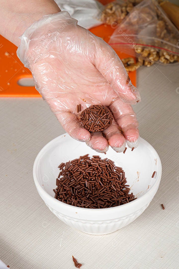 Brigadeiro na mão de um confeiteiro ao lado de um pote de chocolate granulado