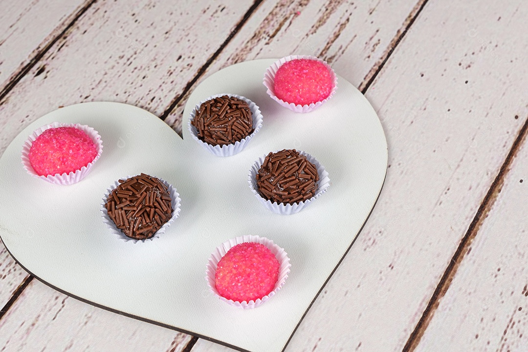 Brigadeiros tradicionais e bichos de pe em dois pratos em forma de coração. Ao lado de um saco de chocolate granulado.