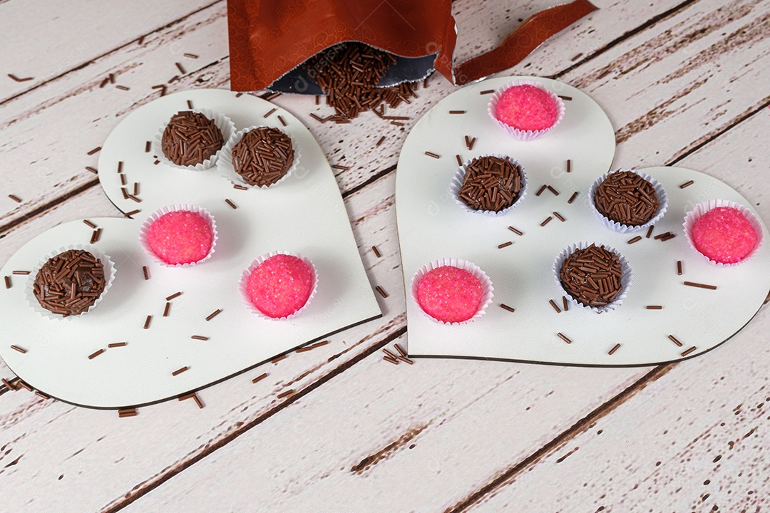 Brigadeiros tradicionais e bichos de pe em dois pratos em forma de coração. Ao lado de um saco de chocolate granulado.