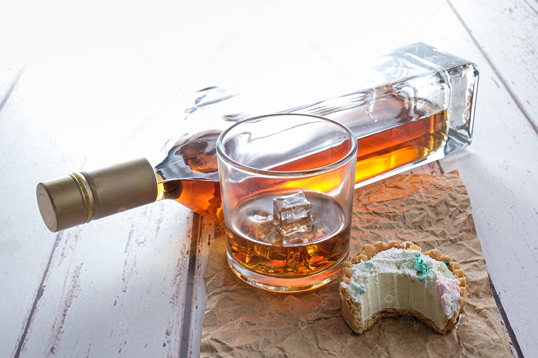 Copo de uísque com gelo em papel pardo, ao lado de uma mini torta