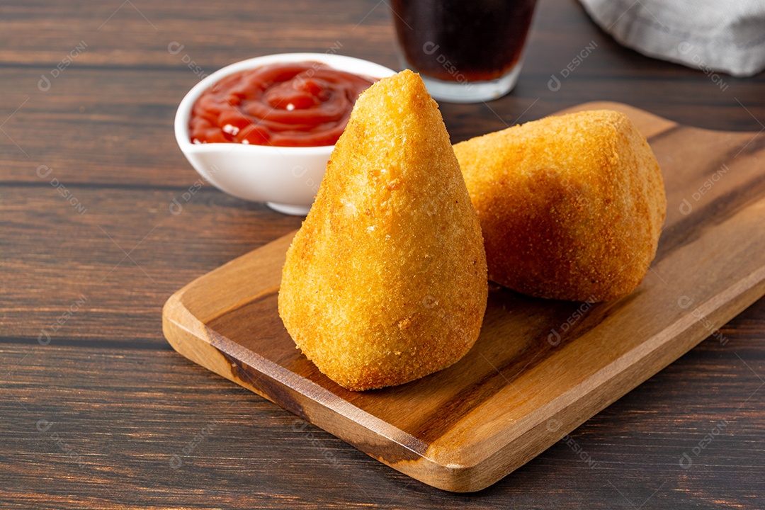 Coxinha lanche tradicional brasileiro frito em óleo e recheado com carne bovina ou carne de frango, com ketchup e refrigerante no fundo sobre mesa de madeira. foto vertical