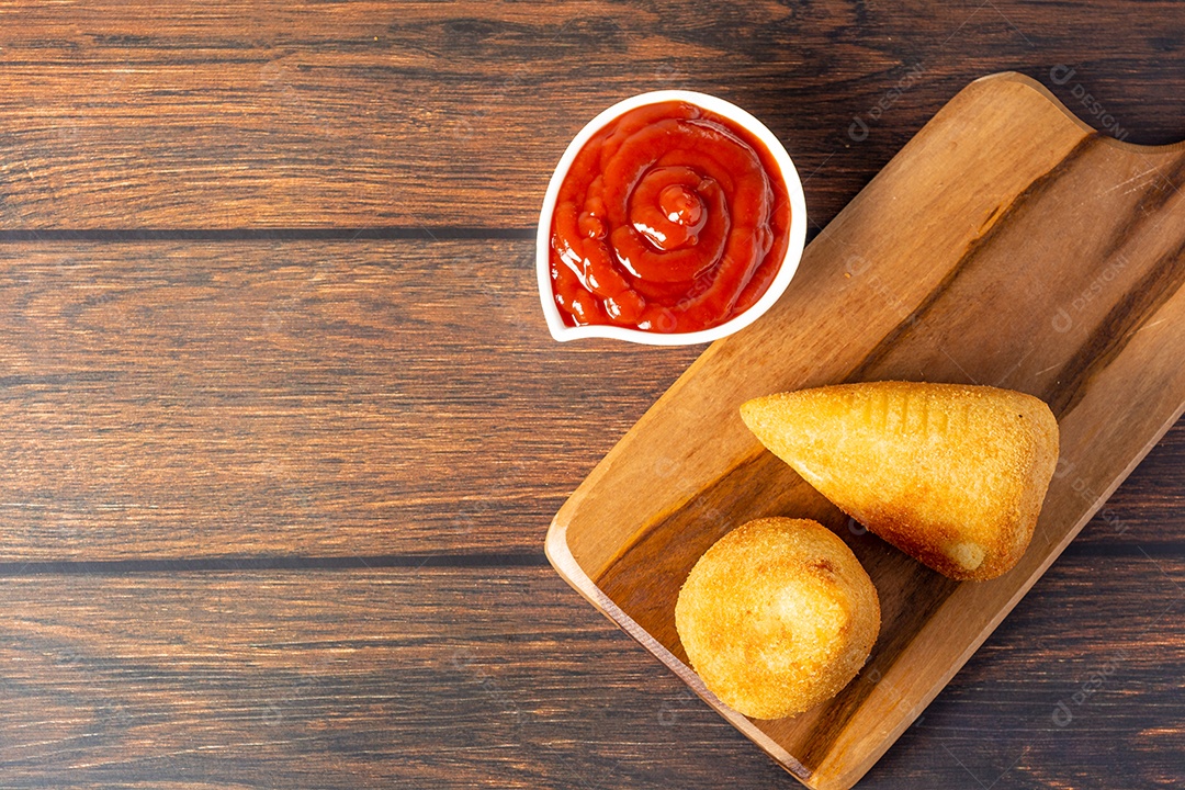 Coxinha lanche tradicional brasileiro frito em óleo e recheado com carne bovina ou carne de frango, com ketchup e refrigerante no fundo sobre mesa de madeira. foto vertical