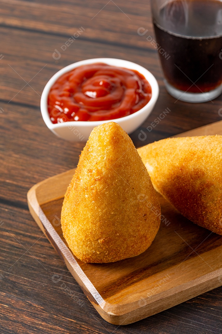 Coxinha lanche tradicional brasileiro frito em óleo e recheado com carne bovina ou carne de frango, com ketchup e refrigerante no fundo sobre mesa de madeira. foto vertical