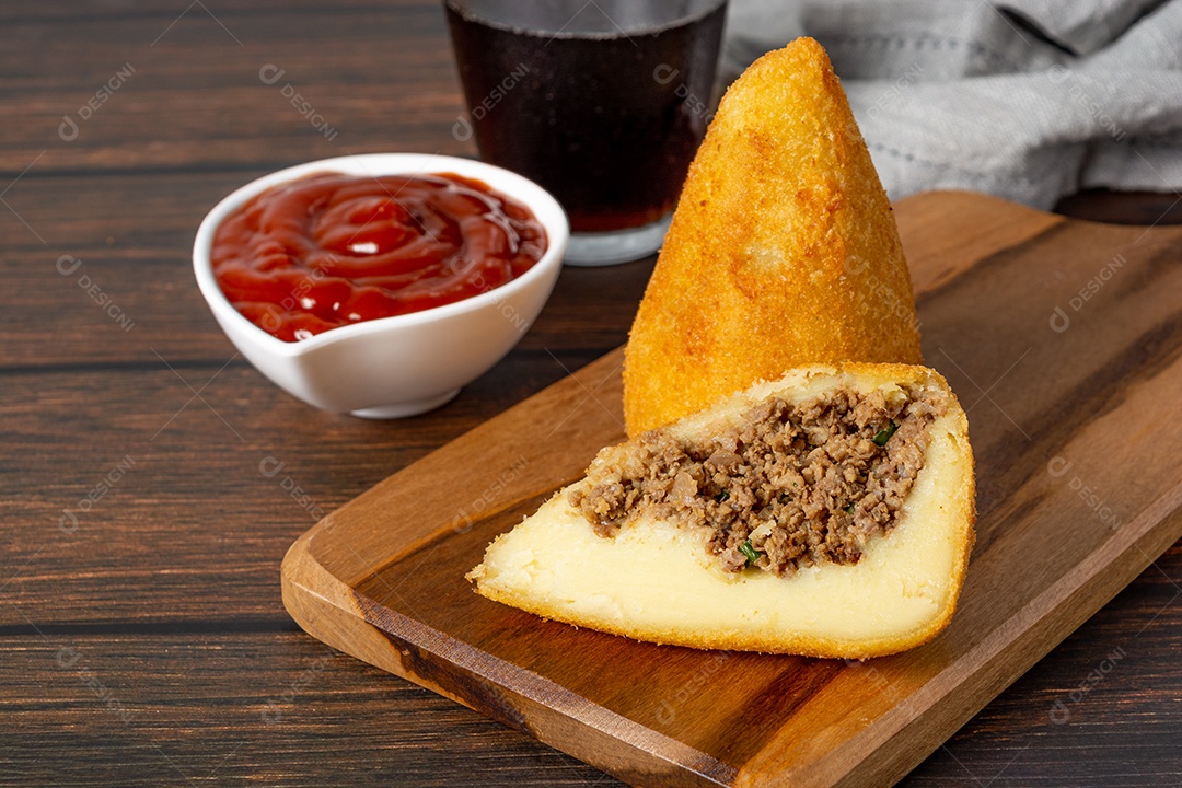 Coxinha lanche tradicional brasileiro frito em óleo e recheado com carne bovina ou carne de frango, com ketchup e refrigerante no fundo sobre mesa de madeira. foto vertical