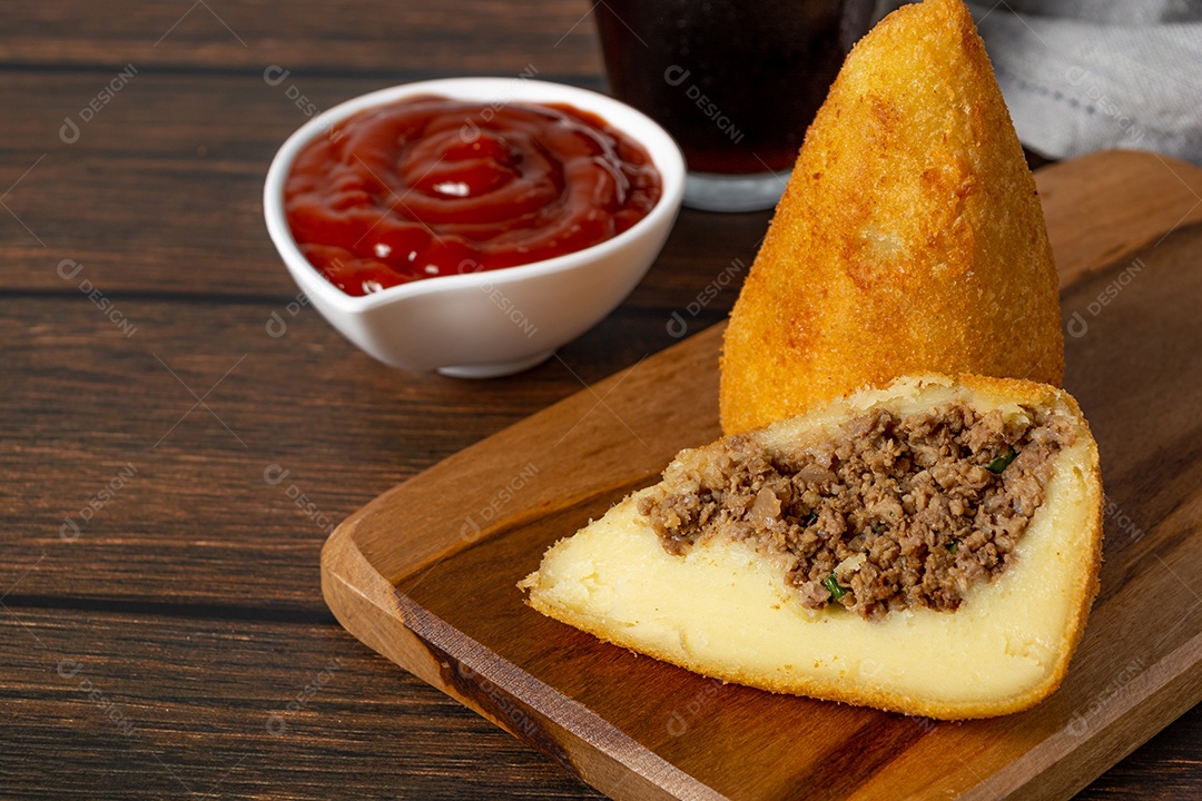 Coxinha lanche tradicional brasileiro frito em óleo e recheado com carne bovina ou carne de frango, com ketchup e refrigerante no fundo sobre mesa de madeira. foto vertical