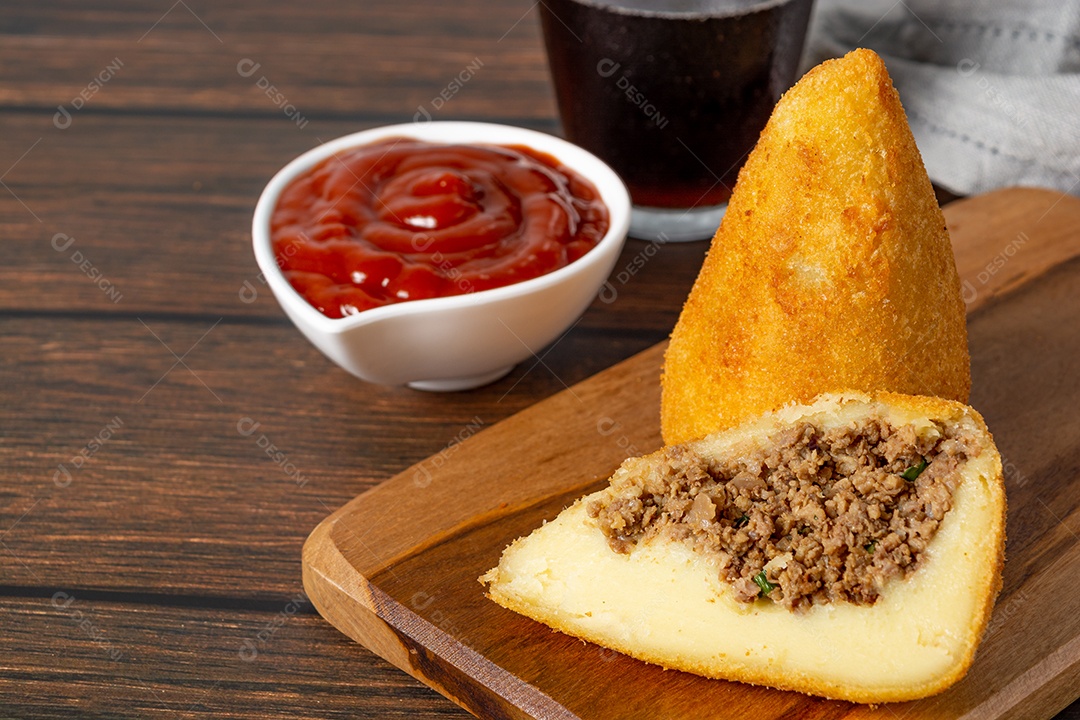 Coxinha lanche tradicional brasileiro frito em óleo e recheado com carne bovina ou carne de frango, com ketchup e refrigerante no fundo sobre mesa de madeira. foto vertical