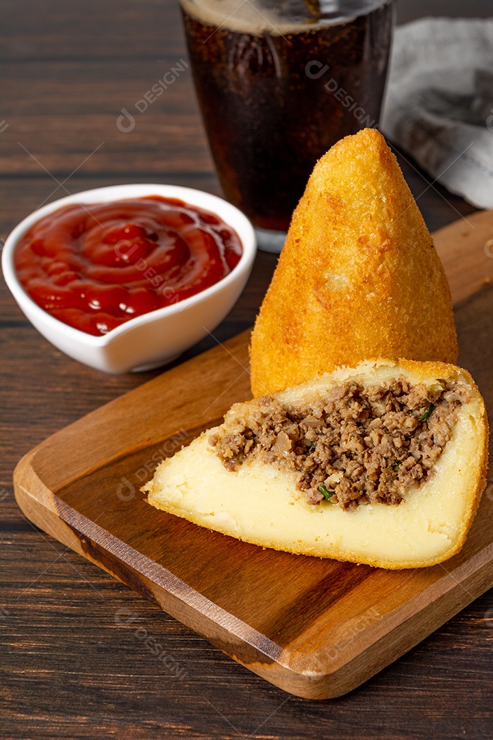 Coxinha lanche tradicional brasileiro frito em óleo e recheado com carne bovina ou carne de frango, com ketchup e refrigerante no fundo sobre mesa de madeira. foto vertical