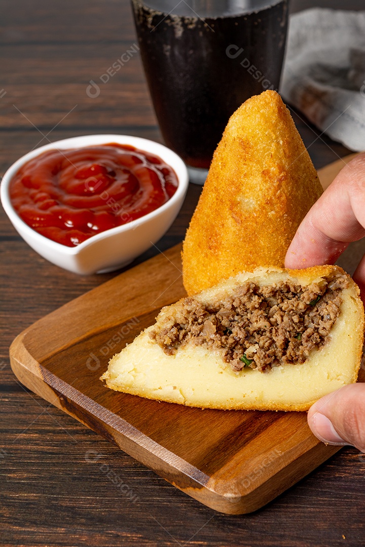 Coxinha lanche tradicional brasileiro frito em óleo e recheado com carne bovina ou carne de frango, com ketchup e refrigerante no fundo sobre mesa de madeira. foto vertical