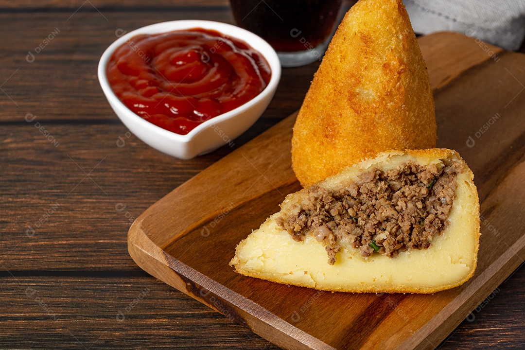 Coxinha lanche tradicional brasileiro frito em óleo e recheado com carne bovina ou carne de frango, com ketchup e refrigerante no fundo sobre mesa de madeira. foto vertical