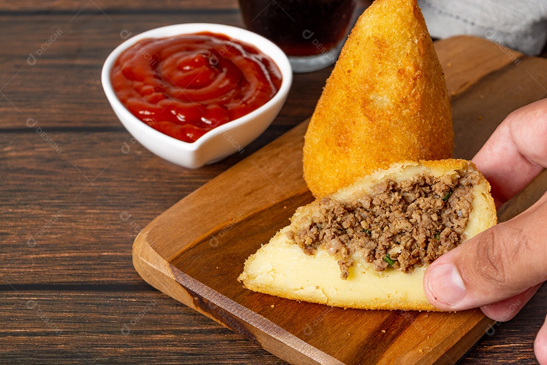 Coxinha lanche tradicional brasileiro frito em óleo e recheado com carne bovina ou carne de frango, com ketchup e refrigerante no fundo sobre mesa de madeira. foto vertical