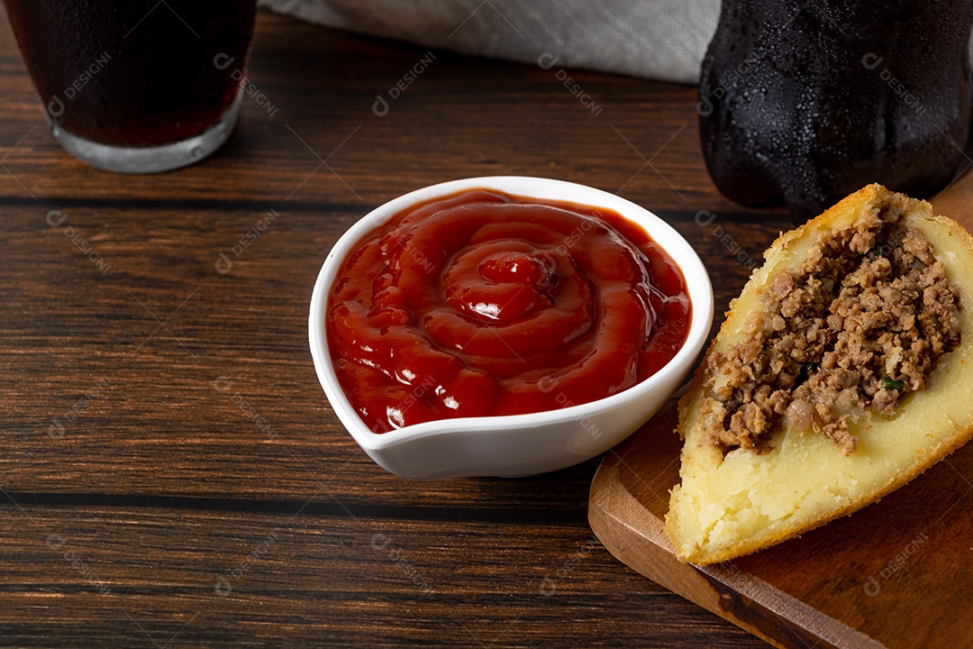 Coxinha lanche tradicional brasileiro frito em óleo e recheado com carne bovina ou carne de frango, com ketchup e refrigerante no fundo sobre mesa de madeira. foto vertical