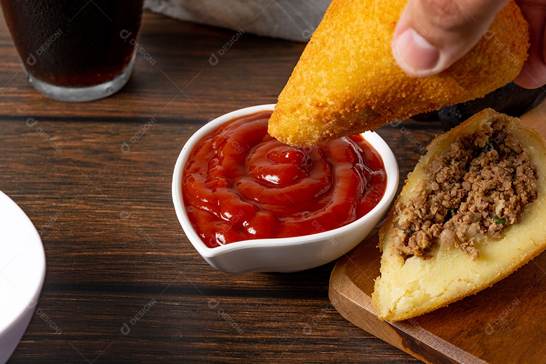Coxinha lanche tradicional brasileiro frito em óleo e recheado com carne bovina ou carne de frango, com ketchup e refrigerante no fundo sobre mesa de madeira. foto vertical