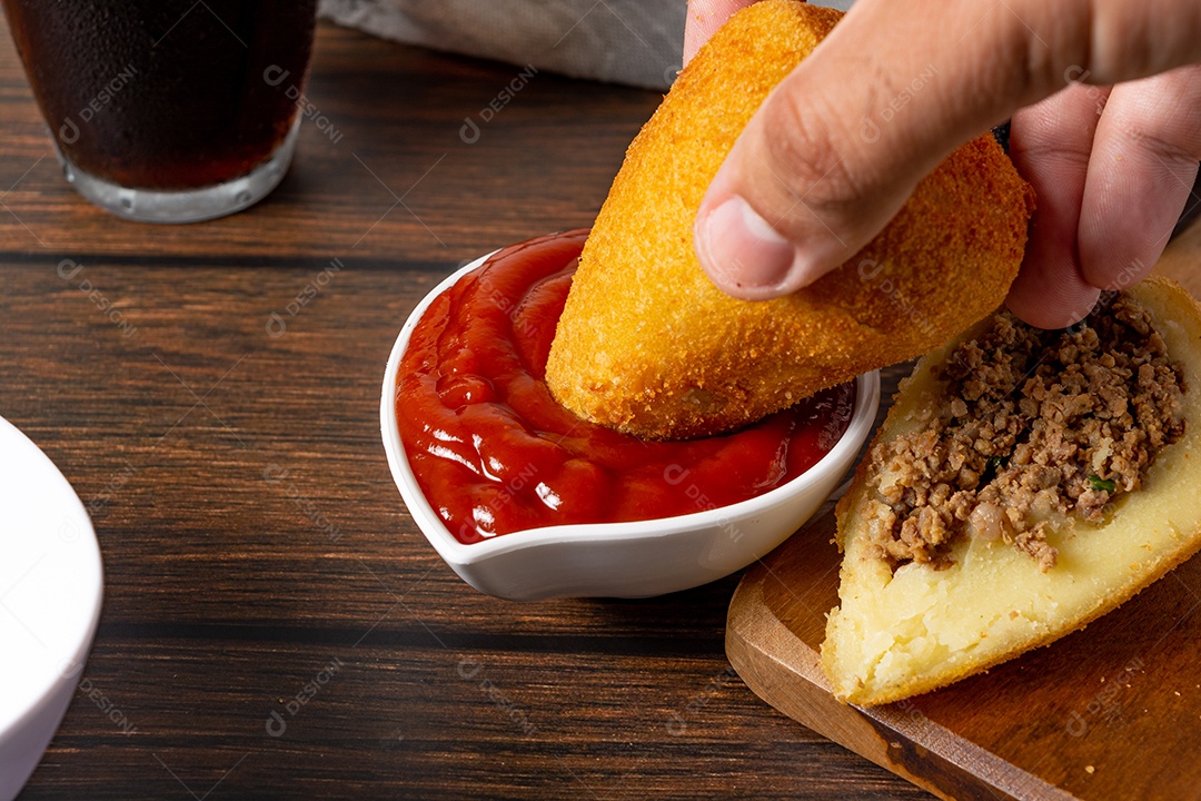 Coxinha lanche tradicional brasileiro frito em óleo e recheado com carne bovina ou carne de frango, com ketchup e refrigerante no fundo sobre mesa de madeira. foto vertical