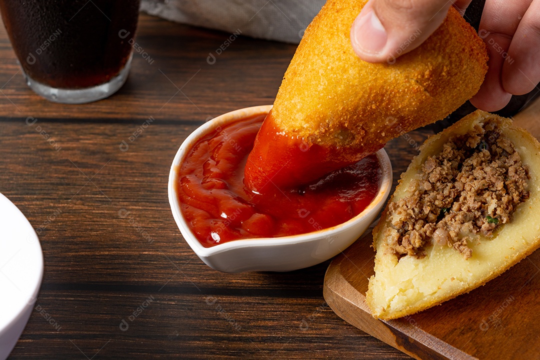 Coxinha lanche tradicional brasileiro frito em óleo e recheado com carne bovina ou carne de frango, com ketchup e refrigerante no fundo sobre mesa de madeira. foto vertical