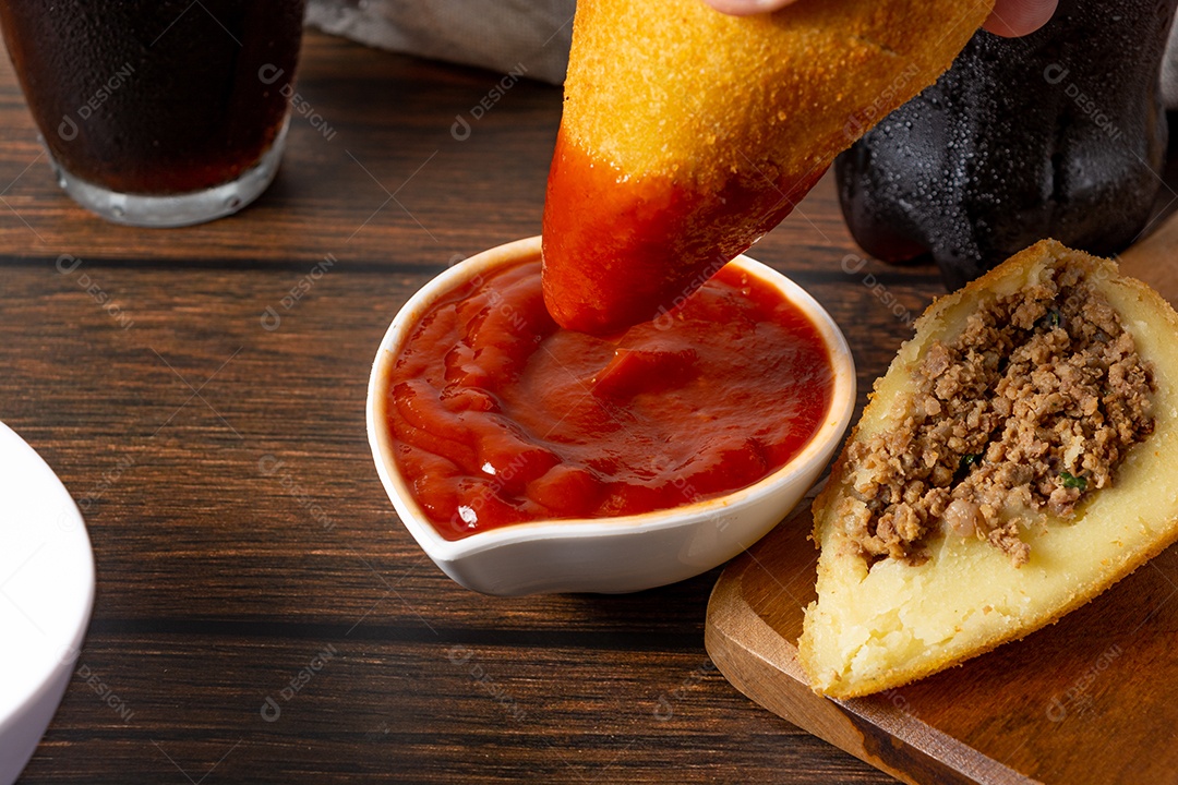 Coxinha lanche tradicional brasileiro frito em óleo e recheado com carne bovina ou carne de frango, com ketchup e refrigerante no fundo sobre mesa de madeira. foto vertical