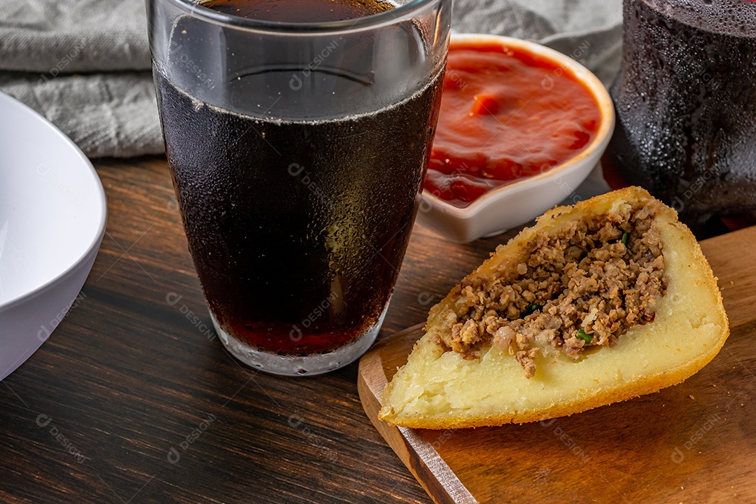 Coxinha lanche tradicional brasileiro frito em óleo e recheado com carne bovina ou carne de frango, com ketchup e refrigerante no fundo sobre mesa de madeira. foto vertical