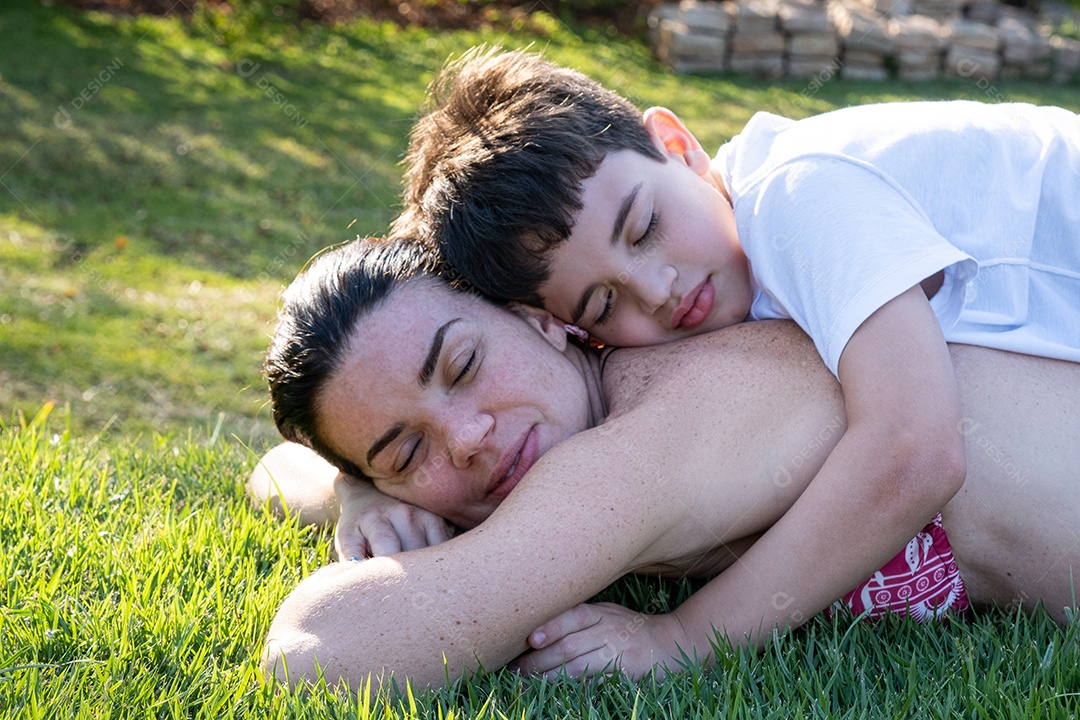 Mãe e filho brasileiros com os olhos fechados deitados no gramado em uma tarde ensolarada