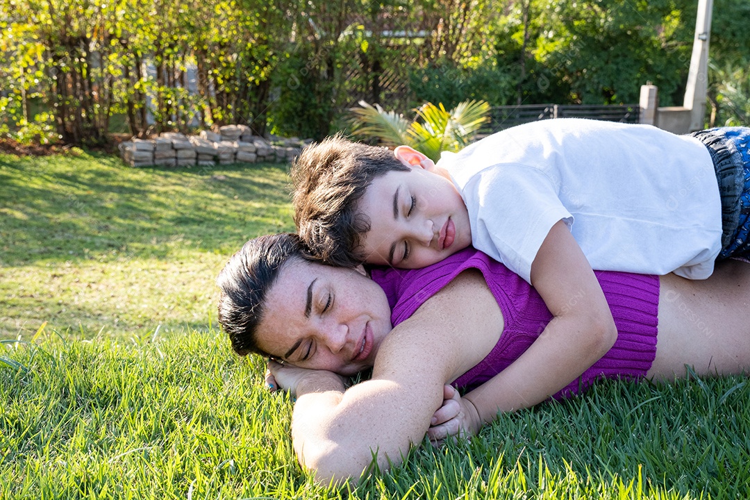 Mãe e filho brasileiros com os olhos fechados deitados no gramado em uma tarde ensolarada