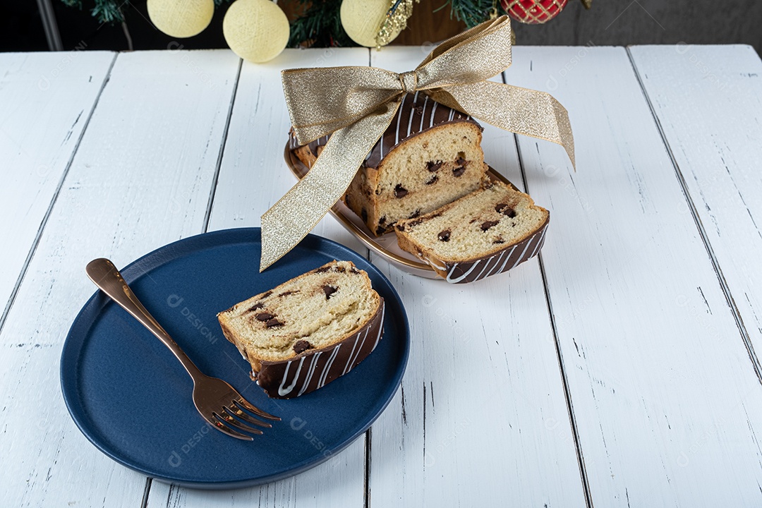 Fatia de chocotone em um prato, ao lado de um garfo, laço de fita e uma árvore de natal