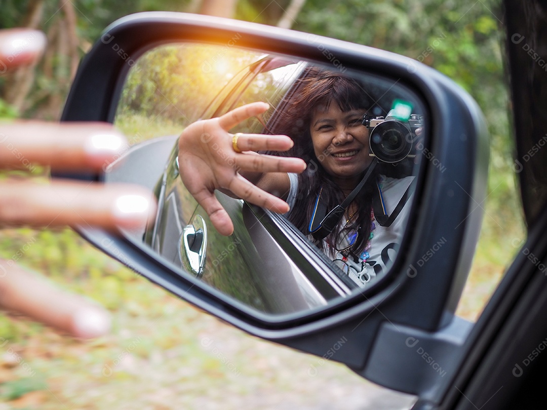 Uma mulher segura uma câmera digital e tira uma foto de si mesma sorrindo refletida no retrovisor do carro.