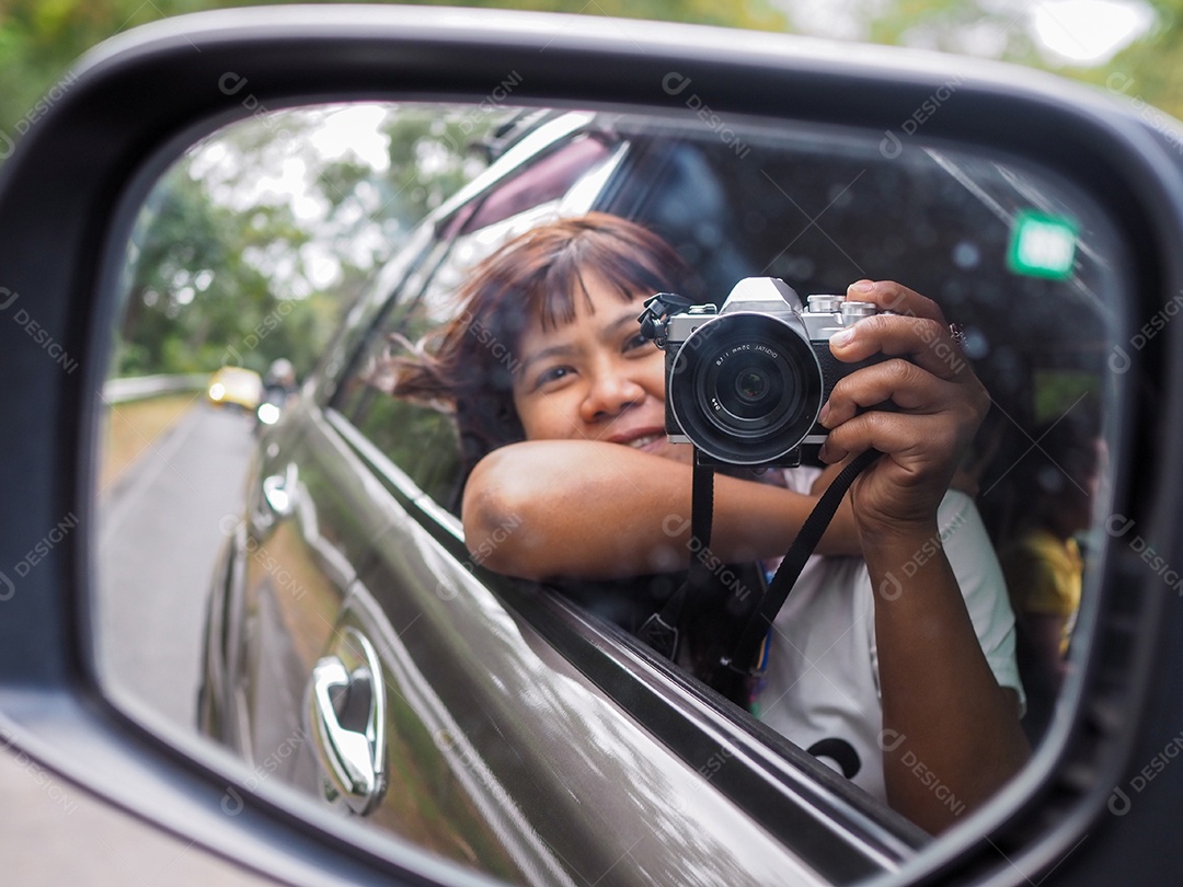 Uma mulher segura uma câmera digital e tira uma foto de si mesma sorrindo refletida no retrovisor do carro.