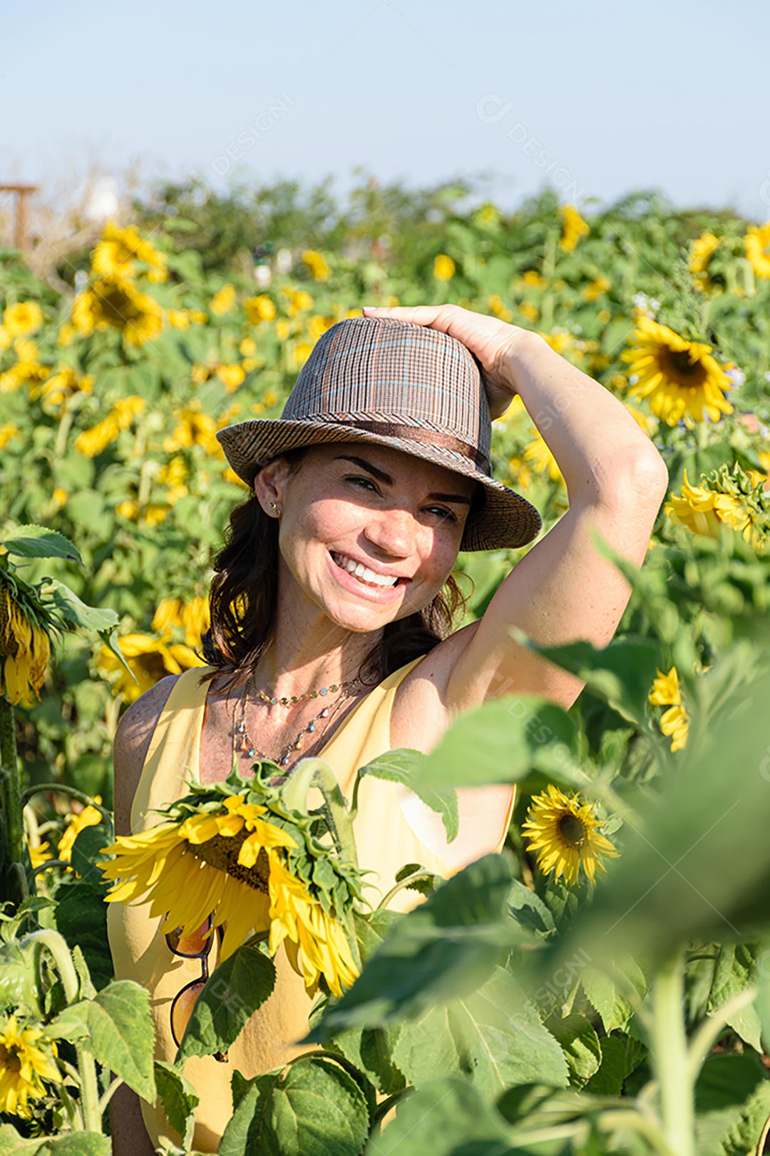 Mulher sorridente em seus 40 anos segurando seu chapéu em um campo de girassol