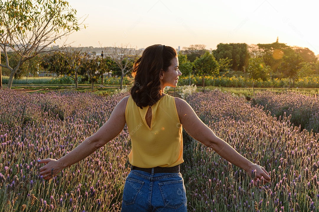 Mulher brasileira de 40 anos e costas, com os braços abertos em uma plantação de lavanda