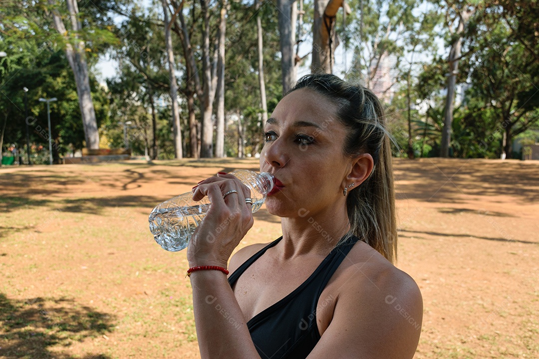 Brasileira de 42 anos, sarada, bebendo água à sombra em praça pública.