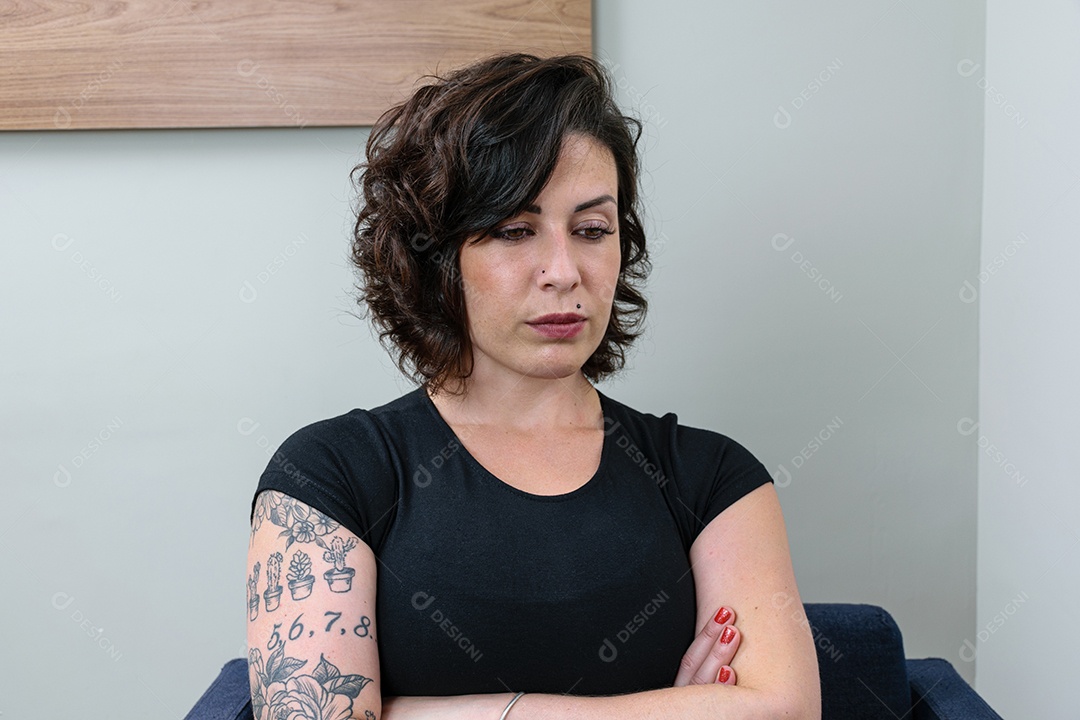 Mulher brasileira, tatuada, com os braços cruzados e olhando para baixo