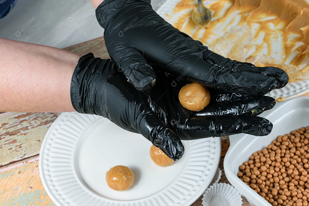 Confeiteiro com luvas pretas fazendo brigadeiro de doce de leite.