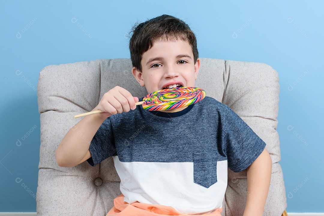 Brasileiro de 8 anos sentado na poltrona. Segurando e pronto para morder um pirulito colorido