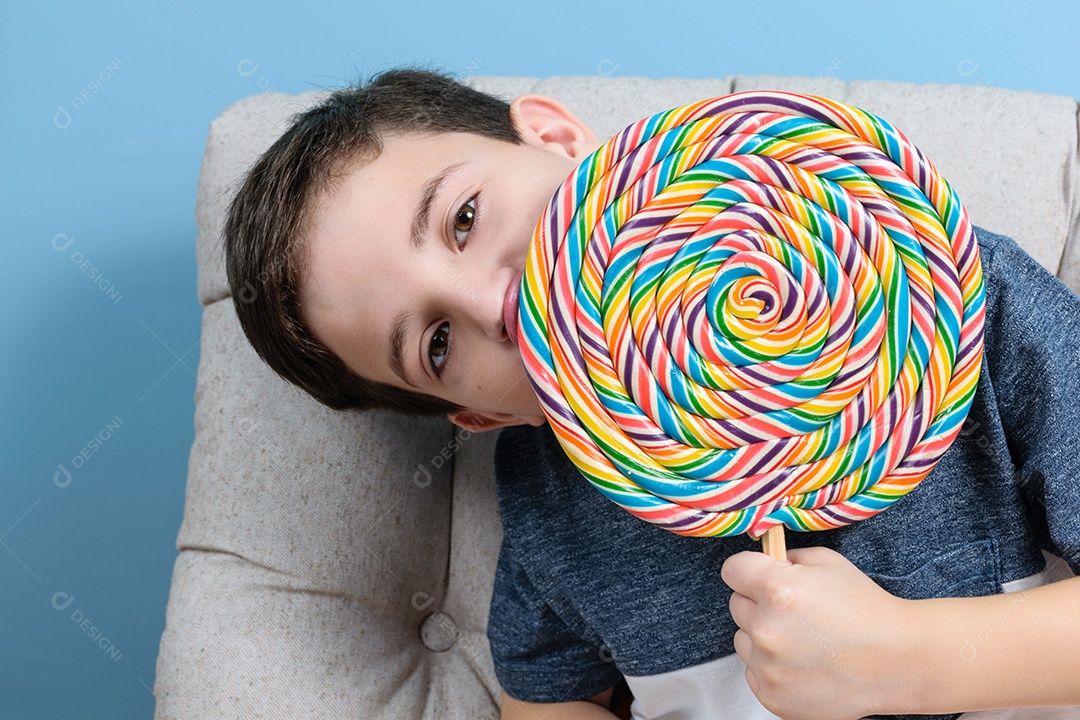 Criança brasileira de 8 anos segurando e lambendo a lateral de um grande pirulito colorido