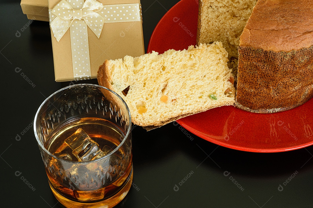 Fatia de panetone em um prato vermelho, ao lado de um copo de uísque