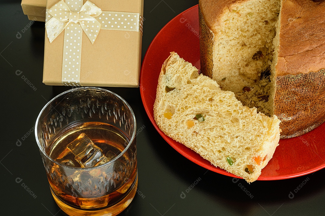 Fatia de panetone em um prato vermelho, ao lado de um copo de uísque